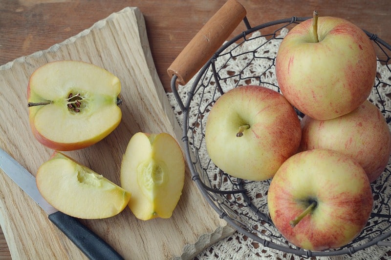 a wire basket of apples next to a cut apple on a cutting board