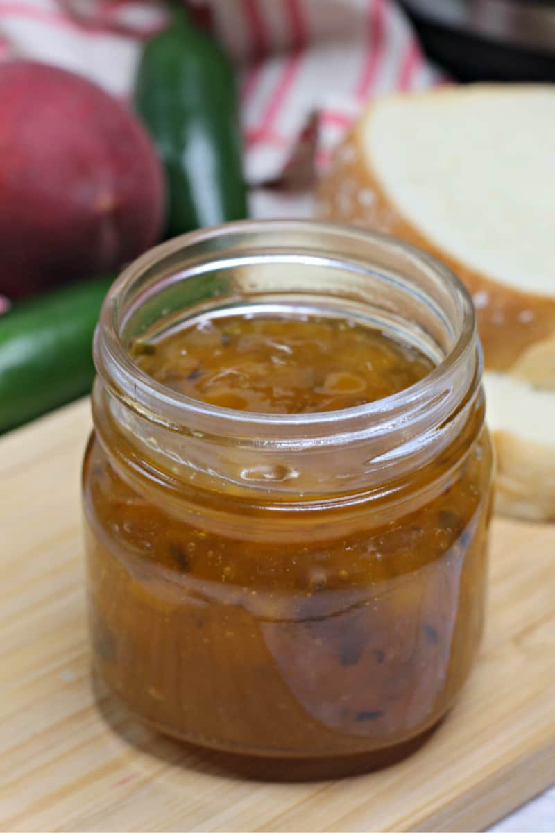 a jar of peach jam with peppers on a wooden cutting board