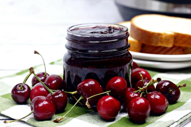 a jar of cherry jam near a pile of cherries on a green checked cloth